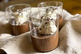 Three servings of chocolate mousse, served in glass tumblers, topped with whipped cream and cacao nibs