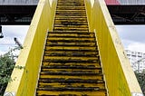 A photo of a steep vertical old, outdoor,  yellow staircase going up towards a bridge.