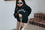 A female child charmingly posing for a picture while wearing sunglasses and holding a bag.