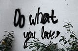 The words “Do what you love!” spray-painted in black on a white wall. Green plants frame the bottom of the image.