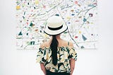 Image of a person with long hair in a ponytail wearing a colorful patterned top (blacks, yellows, some orange) of their shoulders. They are wearing a white hat brimmed with black. They are looking at a colorful splatter abstract painting that almost matches the top.