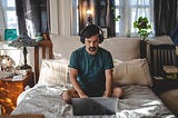 Man in green shirt and wearing headphones sits on his bed working on his laptop from his bedroom.