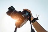 A hand holding an analogue camera in front of the sun with the sky in the background