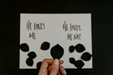 Picture of a white card with He Loves Me, He Loves Me Not written on it. Black petals on each side.