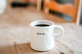 A white mug with the word ‘begin’ on it has a dark drink inside. The mug is sat on top of a light brown wooden table.