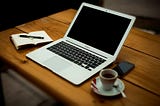 a laptop on a wooden table with a notebook and pen as well as a phone and cup of coffee