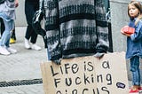 Man in clown makeup including a red-orange wig and a sweater. He has a blank expression on his face and a sign on cardboard that says “Life is a fucking circus.”