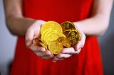 An out of focus image of a lady in a red dress holding to the front of the picture a handful of golden coins which look like chocolate money
