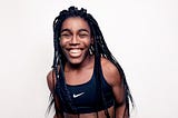 A teenage Black girl with long, thin braids laughs as she stands against a white background. She wears a black Nike cropped running tank.