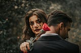 A woman in clown makeup holds a rose while embracing a man who is turned away from the camera.