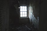 A eight panel, lift up, rectangular wide but square shaped window glowing with incoming light into a darker space impeding seeing outside from the distance the picture is taken. The walls are scrapped and peeling of paint and wall sections; with the left side we are facing exposed brick.