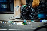 A pile of trash on the sidewalk, some loose litter is strewn in the gutter.