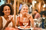 Two girlfriends eating an appetizer in a crowded restaurant. Out of focus, in the background, you can see lots of other people eating in this restaurant.