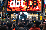 Enthusiastic crowd raising their hands and taking photos with their phones in front of an illuminated billboard in Times Square, featuring the announcement ‘dbt 1.8’ with glowing, festive lights.
