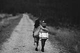 Two young children, one draping an arm over the other, both facing away from the camera, walking down a dirt/gravel road to a distance not established. Upon the sides of the road are brush that are bristlely, short, and dry grass.