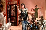 Zombie Eddie dismounts his motorcycle to point an angry finger at something off-camera, while his ex-girlfriend paws at him. Tim Curry in drag watches from the background.