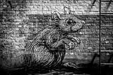 Black and white photograph of a mural line drawing of an angry-looking mouse on a brick wall with a metal ladder leaning against it.