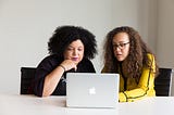 Two women sit at a deskand look at a laptop together.