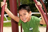 Photo of a small boy smiling on a piece of playground equipment.