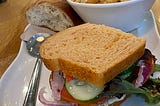 Close-up on a large white plate with a vegetable sandwich next to a bowl of tomato soup topped with croutons and on the other side of the bowl is a roll