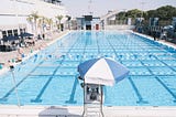 A lap pool with lanes set up short-wise. A lifeguard sits under an umbrella on their tower. Swimmers climb out on the left side. Patrons pepper the pool deck.