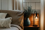 An intimate picture of a bed with neutral bedding beside a nightside table with a lit lamp.