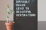 Difficult Roads Lead to Beautiful Destinations — a board bearing this inscription with a seedling in a pot next to it. Hope springs eternal.