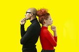 Two people stand back-to-back against a vivid yellow background, each in a thoughtful pose with one hand under the chin. The man on the left wears a black turtleneck and dark glasses, while the woman on the right wears a red top and clear glasses. Their contrasting outfits and mirrored stances create a striking, symmetrical composition.