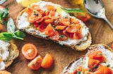 Two slice of bruschetta with cream cheese and tomato sit on a bread board next to a bottle of olive oil