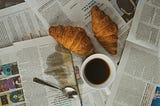Cover image: Many newspapers with a 2 croissants, a spoon, a cup of coffee, and some coffee split over a newspaper.