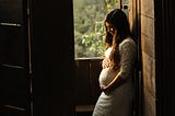 A pregnant woman in a white maternity dress, hands clasped over her belly and looking down admiringly at the new life inside her.