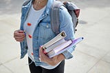 Student on the sidewalk with books on her arm and headphones in her ears.