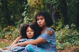 A Black mother and daughter are sitting on the ground in a forest. The daughter is leaning backwards on her mother. They both have serious looks on their faces.