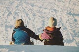 Two young children holding hands sitting on the snow