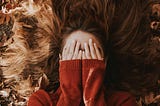 Girl on a floor of dry leaves with her hands over her face and wearing a red jumper