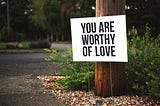 A big white sign that says, “You Are Worthy Of Love”, sits in front of a tree, behind which there is a green bush.