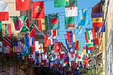 Many of the flags of the world hanging from wires or a clothesline across two buildings. Very colorful.