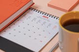 Paper calendar open to January 2024. It is on a desk with a pen, cup of coffee, and notepad