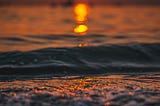 The image shows a vibrant sunset over the ocean. The sun, a glowing orange circle, is low on the horizon, casting a warm, golden reflection on the surface of the water. The waves are gently rippling, with the sunlight creating a shimmering path across them. The overall scene is serene and calming, with the soft, warm colors evoking a peaceful evening by the sea.