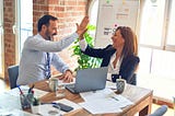 Man and a woman at a desk high-five each other. Computer on desk.