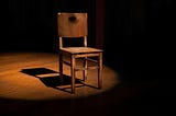 A wooden chair sits on a wooden floor, lit up by a spotlight.