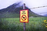 A barbed wire fence in front of a steep hill with some cloud cover at the top. The sign on the fence says “NO Drones.” There are some low buildings at the foot of the hill.