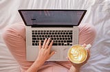 Woman sitting on her bed with her legs crossed drinking coffee and using her laptop.