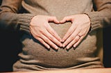 The picture shows a woman in brown sweater forming a heart shape with her hands over the bulge of her tummy.