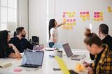 4 people sitting around a table working and a woman pointing to post-its on the wall