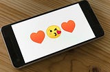 The image shows a smartphone lying on a wooden table, displaying a simple white screen with two red heart emojis and a yellow face emoji blowing a heart kiss. The emoji sequence, featuring playful affection, is centered on the screen, suggesting a message of love or flirtation. The phone is modern, with a black frame and visible buttons, resting in a horizontal position.