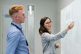 A bussiness woman explaining something to a businessman on a whiteboard