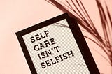 a black and white letterboard spells out “self care isn’t selfish” on a light pink background