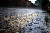A picture of a road in the rain. The image is a close-up near the curb, over a double-yellow line. The rain is beating down heavily and the splashes are clearly visible. Blurred houses are in the background.