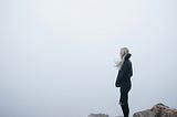 A woman standing on the edge of a rocky cliff, looking off into the foggy drop below